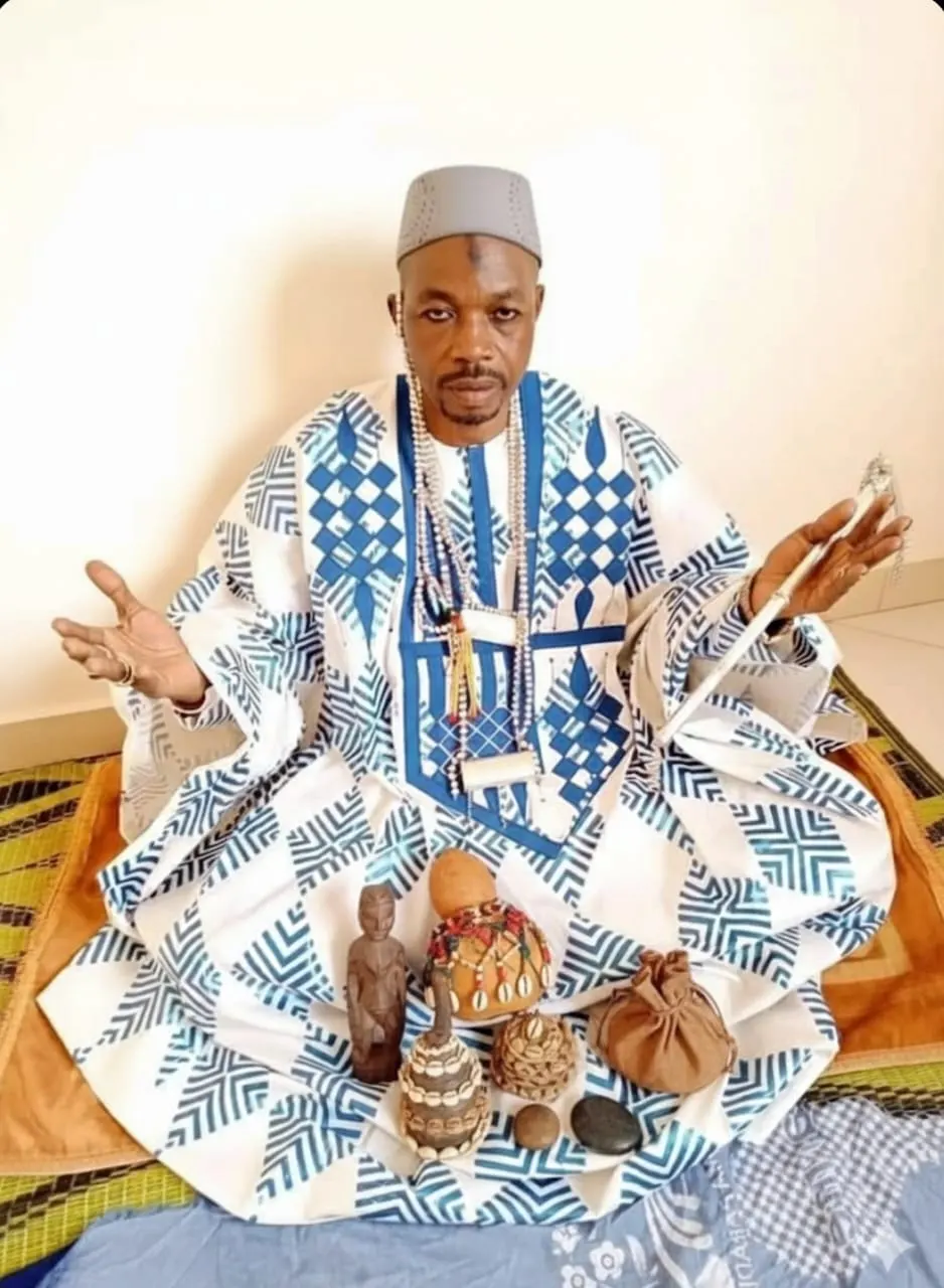 Ndiaye Salifou Malick assis sur une natte, en boubou, devant une calebasse ornée de cauris