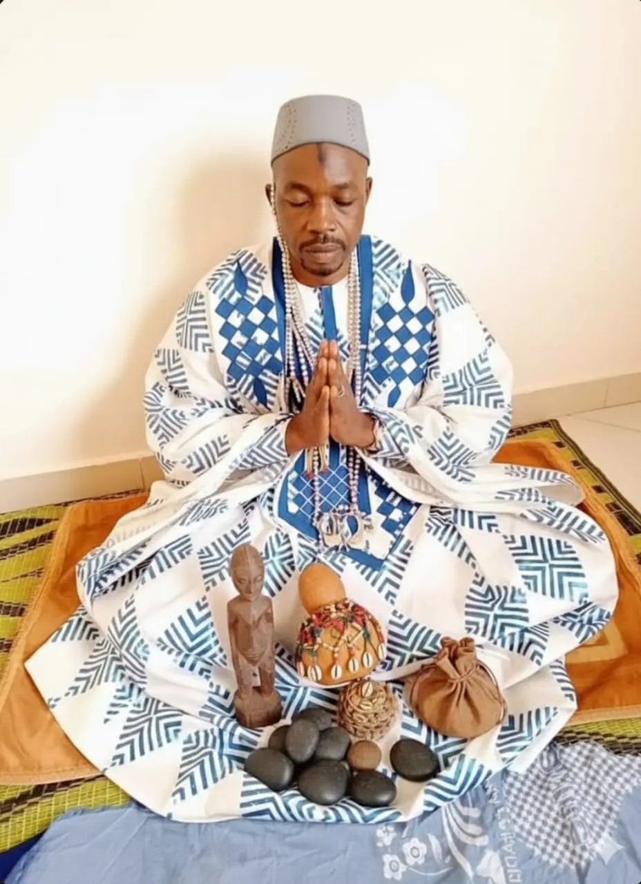 Ndiaye Salifou Malick en boubou blanc et indigo, mains jointes, devant ses objets de culte