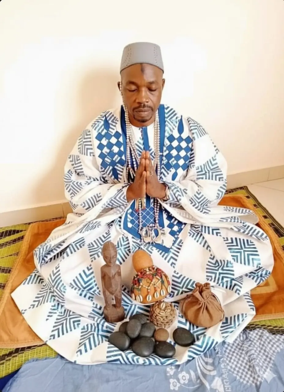Ndiaye Salifou Malick en boubou blanc et indigo, mains jointes, devant ses objets de culte
